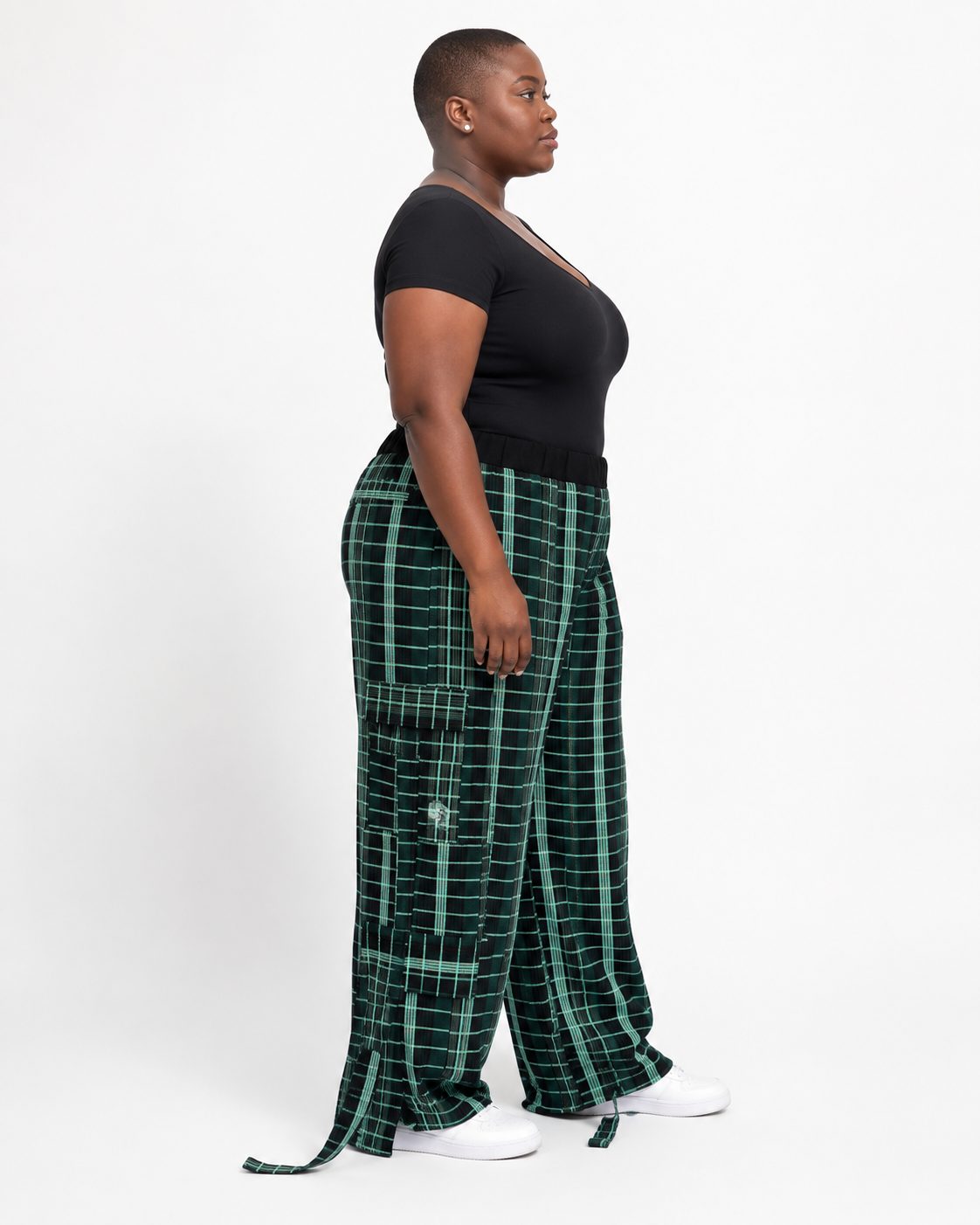 A model shown from the side profile wearing dark green wide-leg plaid pants with a black short-sleeve top and white sneakers, photographed against a plain white studio background.