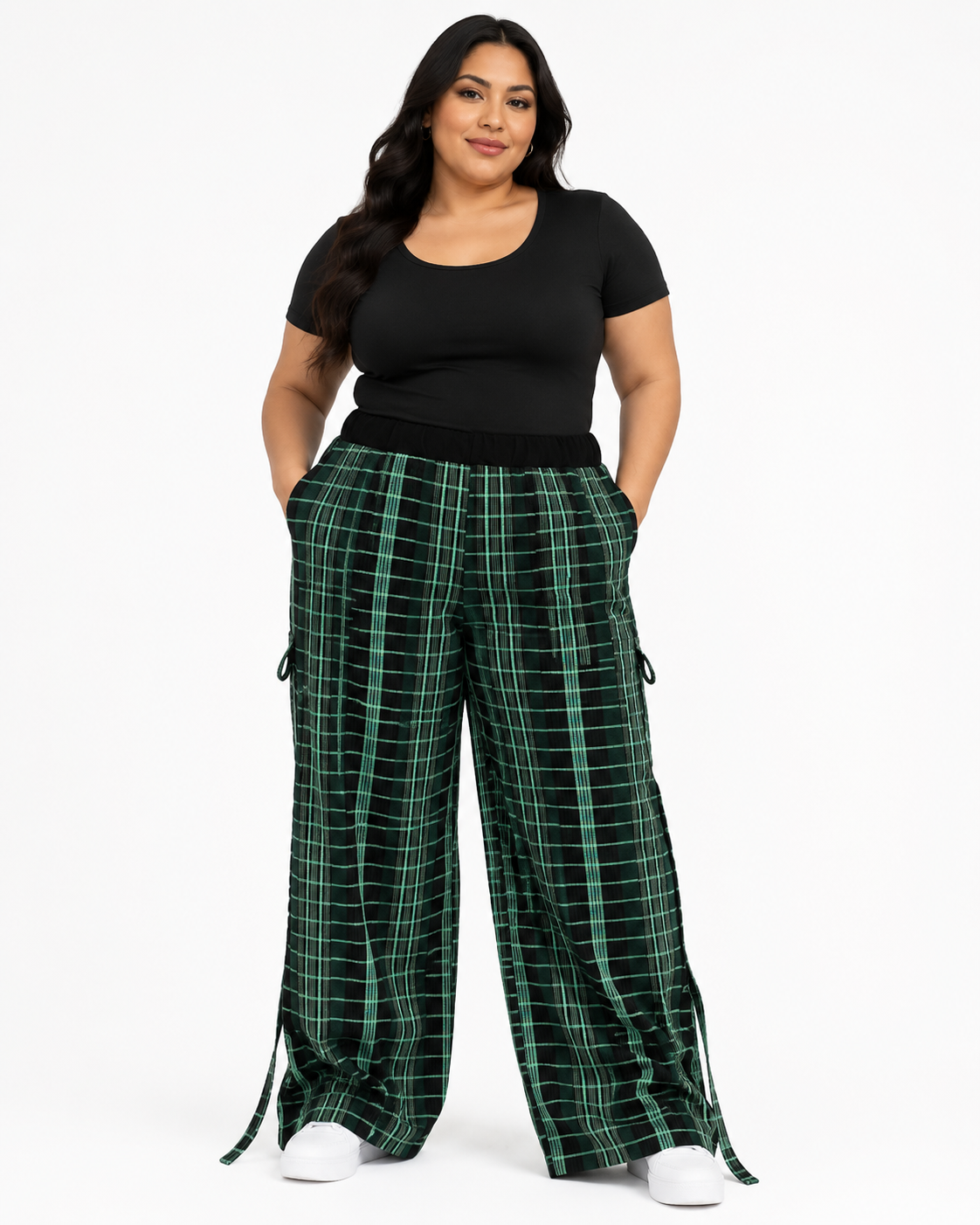 A woman modeling wide-leg green plaid pants with a black scoop-neck top and white sneakers, standing against a plain white studio background.