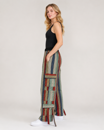 A woman stands in profile wearing a black sleeveless top and very wide-leg, patchwork-style pants with vertical stripes and geometric blocks in muted green, beige, red, and blue. She is wearing white sneakers against a plain light gray studio background.