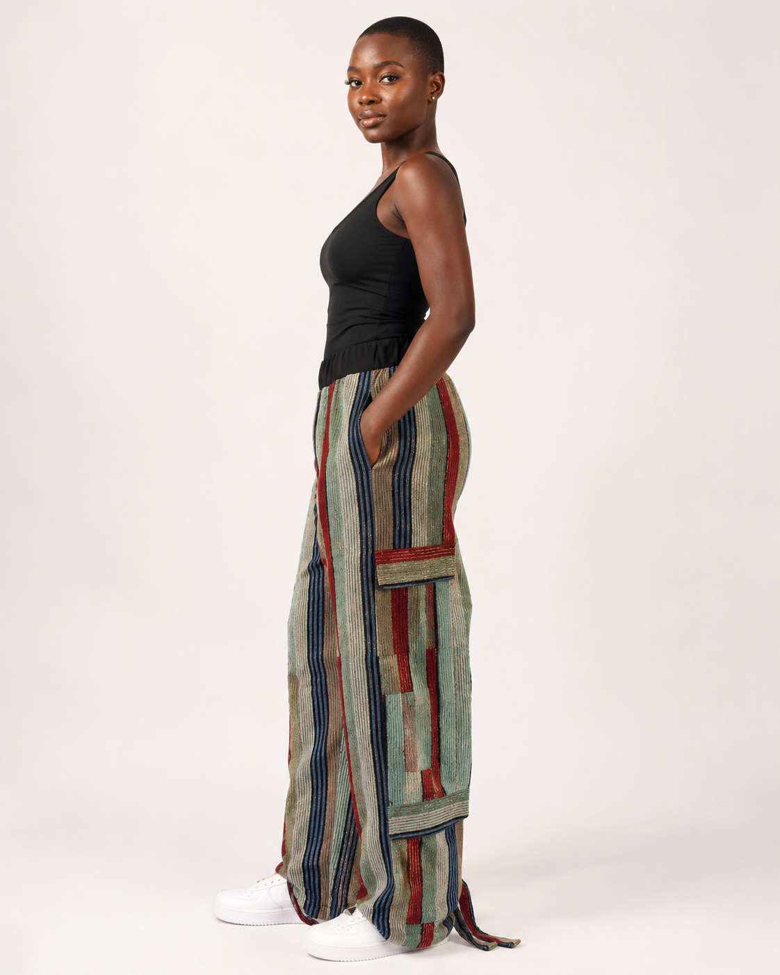 A woman stands in profile wearing a black sleeveless top and wide-leg striped pants with vertical panels in muted green, beige, navy, and rust red, paired with white sneakers, against a plain light background.