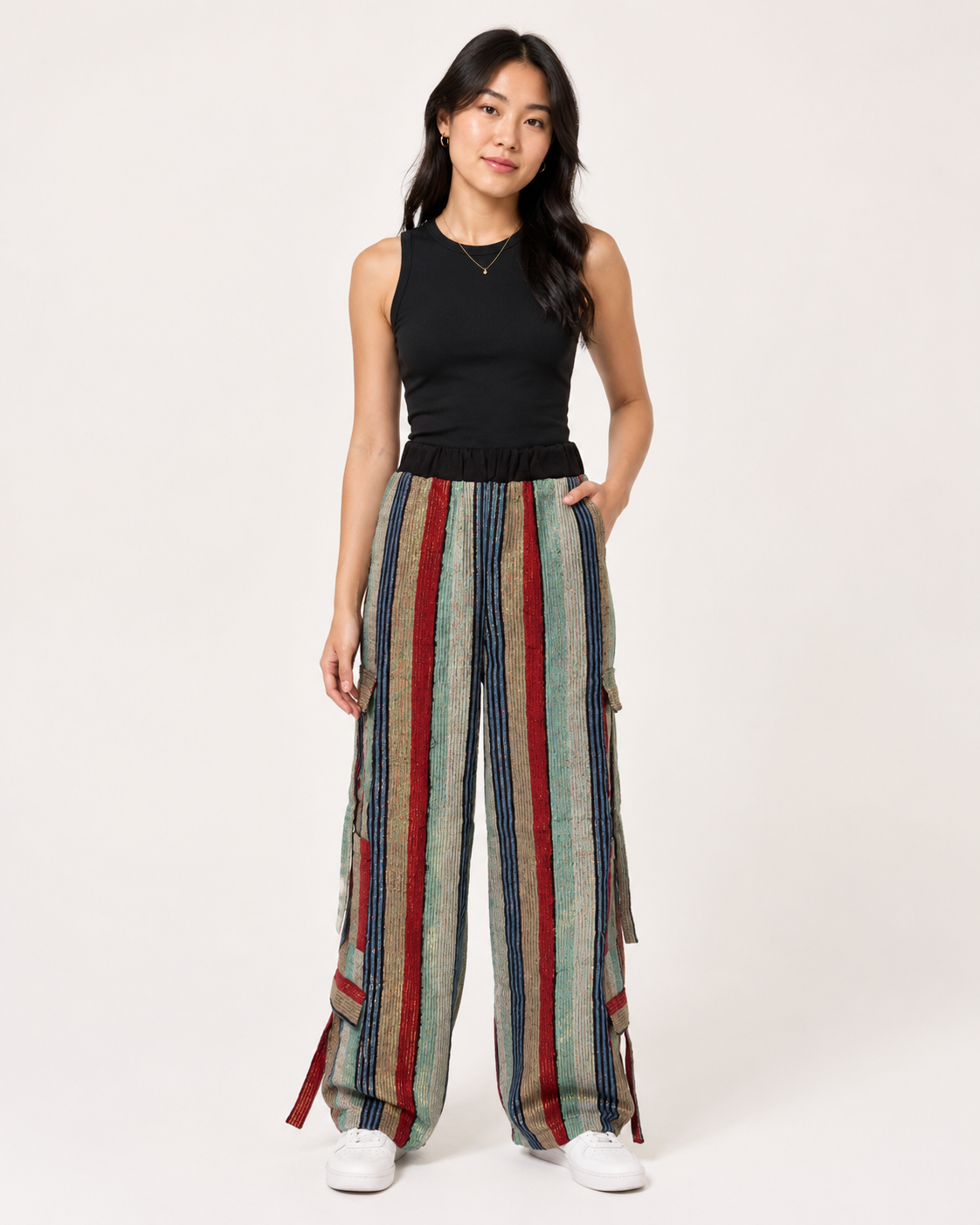 A woman modeling a sleeveless black top and wide-leg striped pants with vertical red, blue, and beige stripes, paired with white sneakers, against a plain light background.