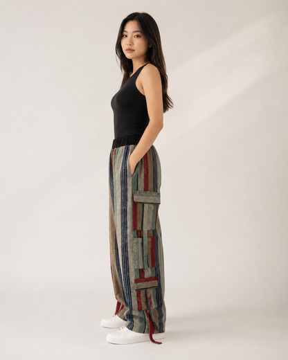 A woman stands in profile wearing a black sleeveless top and wide-leg cargo pants with a multicolor striped and patchwork pattern in blue, red, beige, and gray. She is wearing white sneakers against a plain light beige studio background.