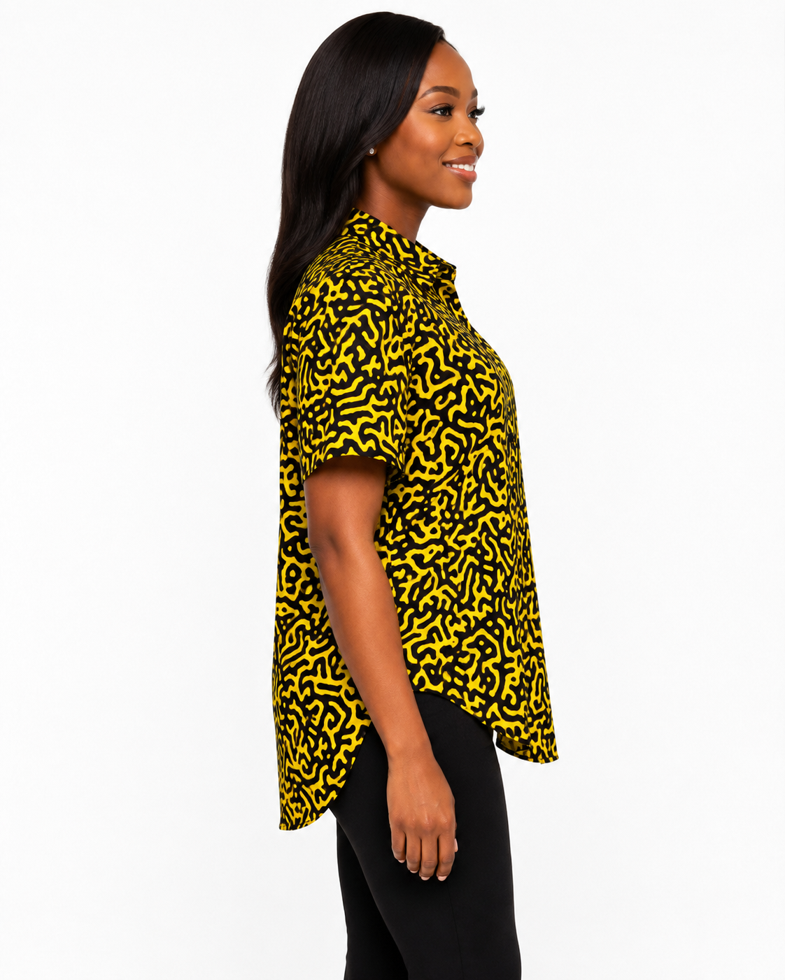 A woman shown in side profile wearing a short-sleeve button-up shirt with a bold yellow and black abstract maze-like pattern, paired with black pants, against a plain white studio background.