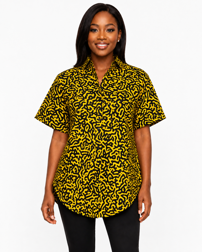 A woman modeling a short-sleeve button-up shirt with a bold yellow and black abstract pattern, paired with black pants, against a plain white studio background.