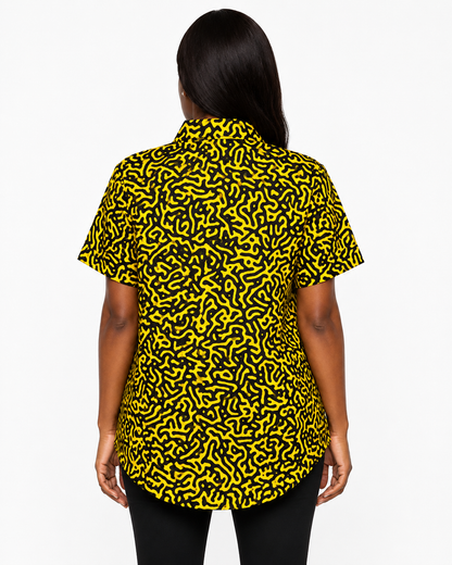 A woman viewed from the back wearing a short-sleeve button-up shirt with a bold black and yellow abstract maze-like pattern, paired with black pants, against a plain white studio background.