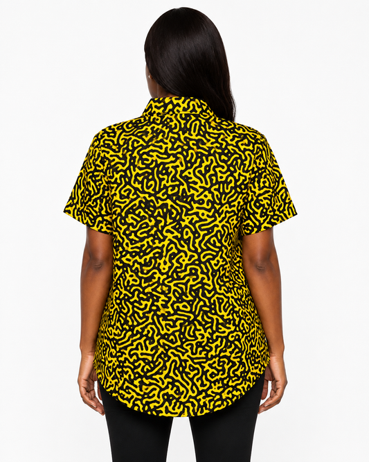 A woman viewed from the back wearing a short-sleeve button-up shirt with a bold black and yellow abstract maze-like pattern, paired with black pants, against a plain white studio background.
