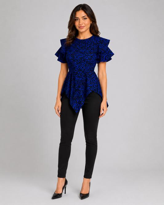 A woman modeling a royal blue short-sleeve top with a black abstract pattern and ruffled shoulders, paired with black skinny pants and black heels, against a light gray studio background.