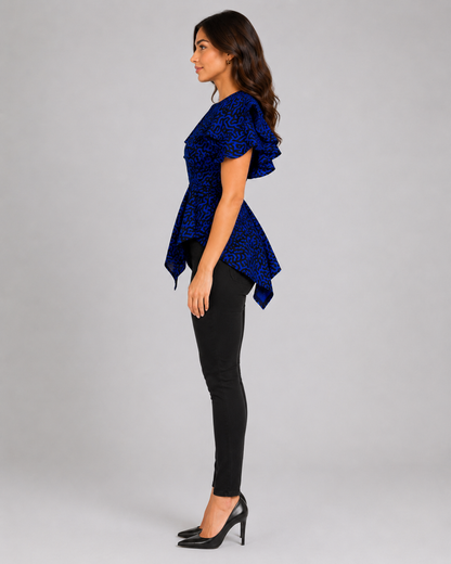 A woman in profile wearing a royal blue patterned peplum top with ruffled sleeves, black skinny pants, and black high heels, standing against a light gray studio background.