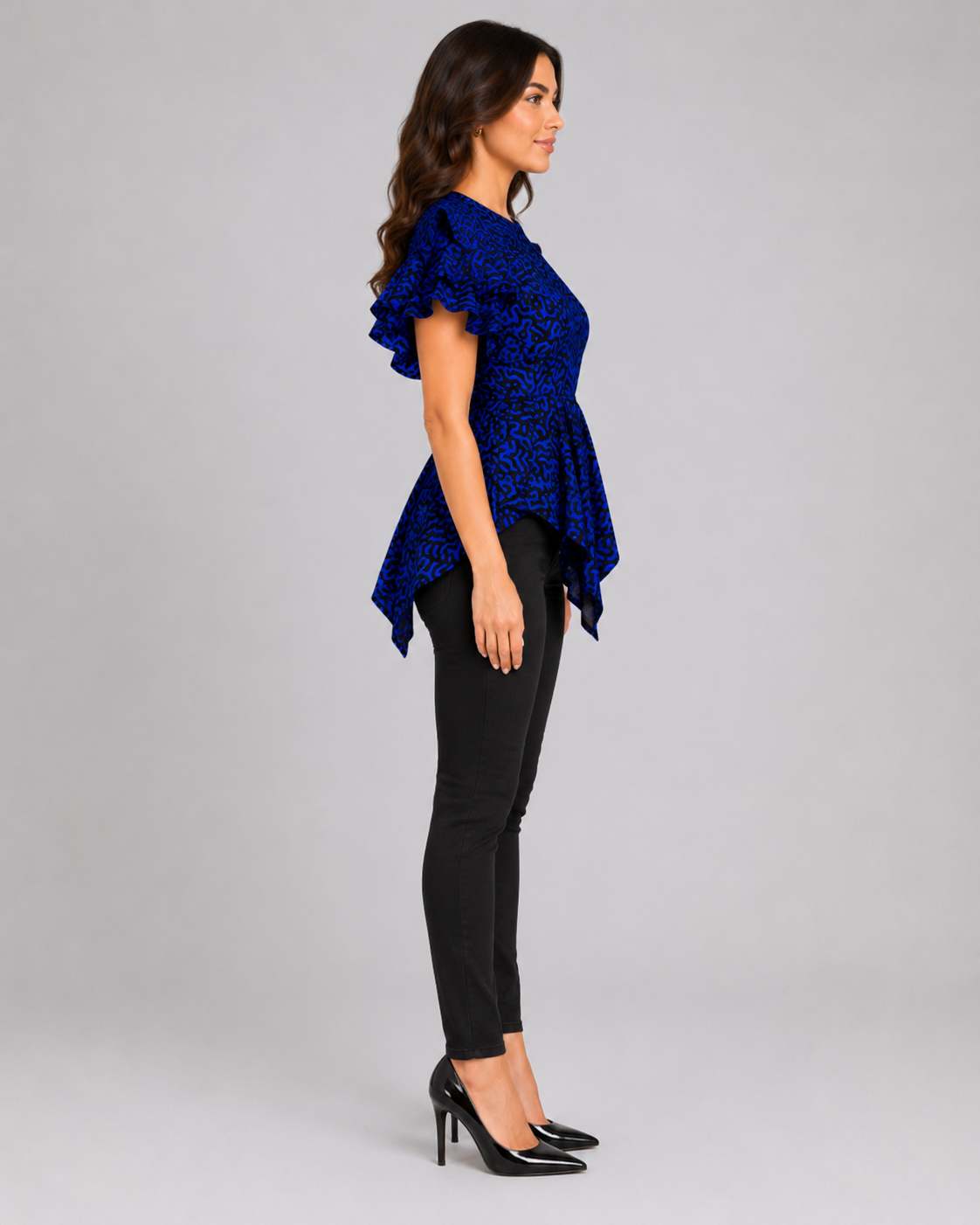 A woman shown in side profile wearing a royal blue patterned ruffled short-sleeve top, black pants, and black heels against a light gray studio background.