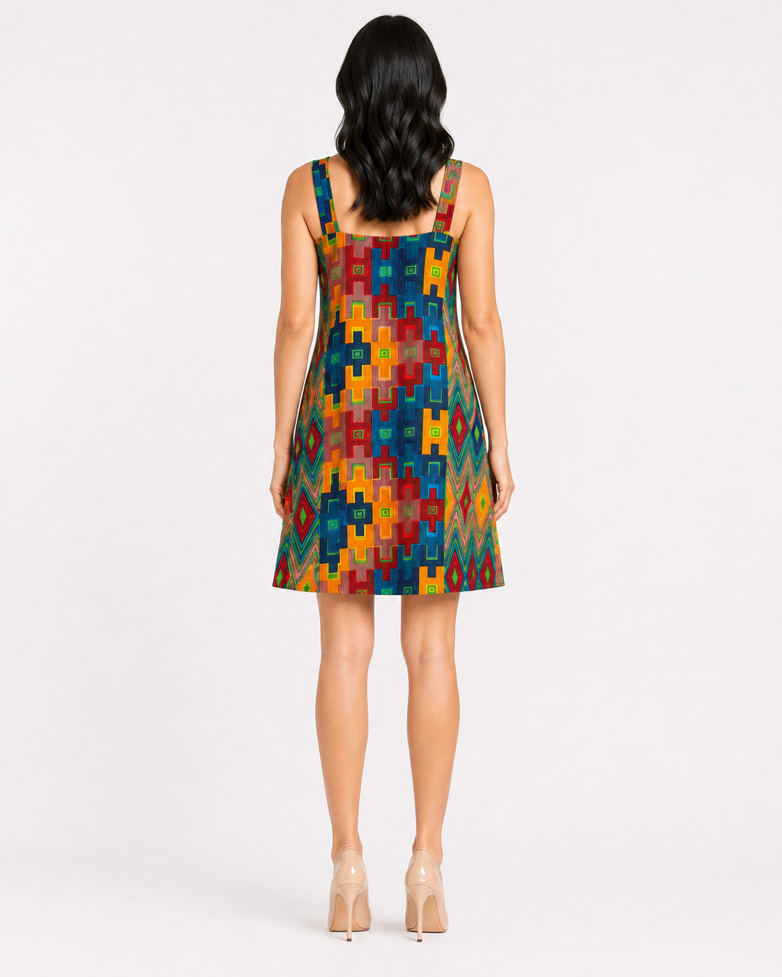 A woman viewed from the back wearing a sleeveless A-line dress with a colorful geometric block pattern in red, yellow, blue, and green, paired with beige high heels against a light gray studio background.