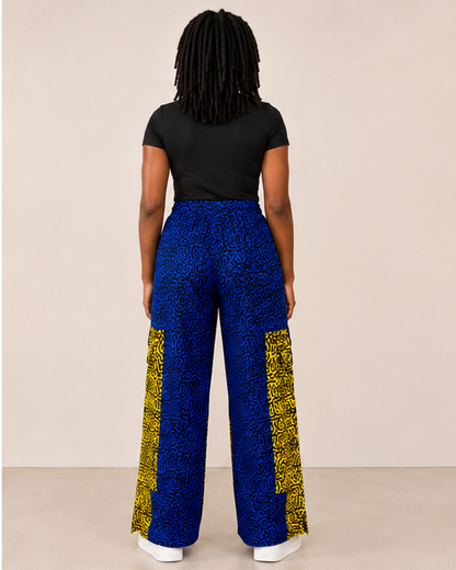 A model shown from the back wearing high-waisted, wide-leg trousers with a blue and black abstract pattern and contrasting yellow side panels, paired with a black t-shirt and white sneakers.