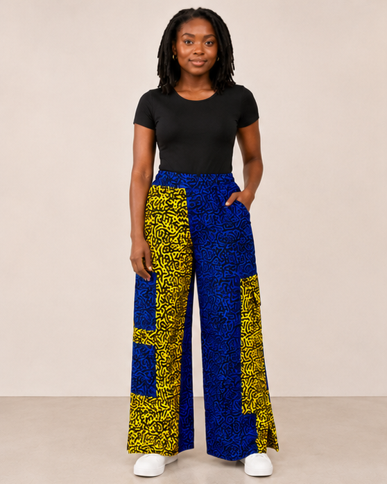 A woman modeling high-waisted, wide-leg pants with a bold blue and yellow patterned design, paired with a black short-sleeve top and white sneakers, against a plain beige studio background.