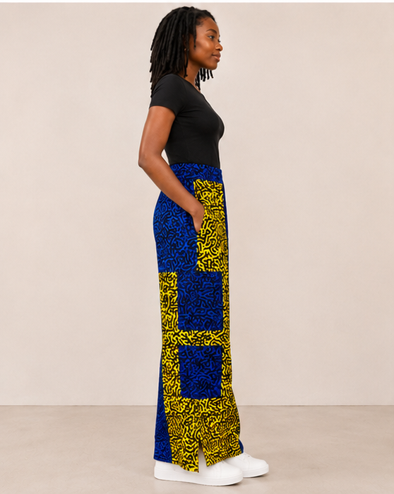 A woman in profile wearing a black short-sleeve top and a long, wide-leg skirt with a bold blue and yellow patterned design, standing against a plain beige studio background.