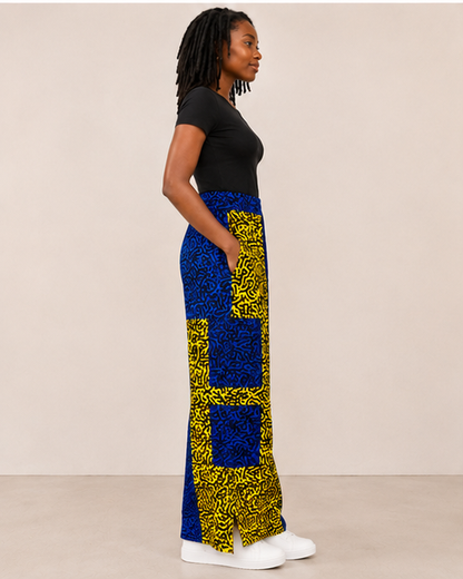 A woman in profile wearing a black short-sleeve top and a long, wide-leg skirt with a bold blue and yellow patterned design, standing against a plain beige studio background.