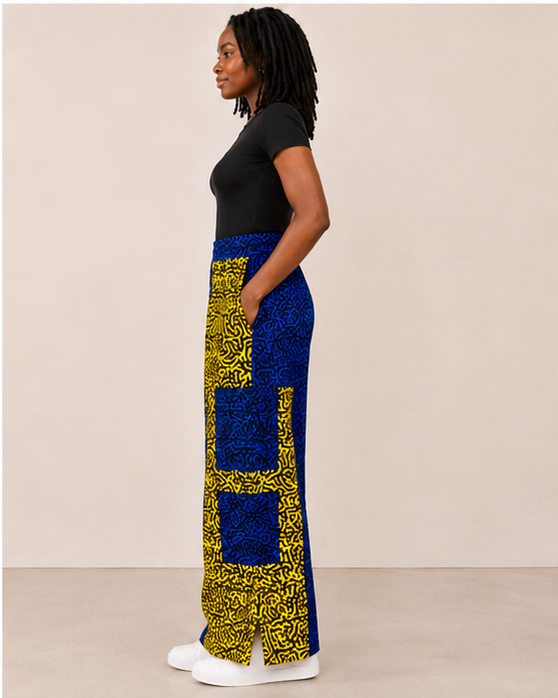 A woman stands in profile wearing a black short-sleeve top and a long, wide-leg skirt with a bold blue and yellow patterned design, paired with white sneakers, against a plain beige studio background.